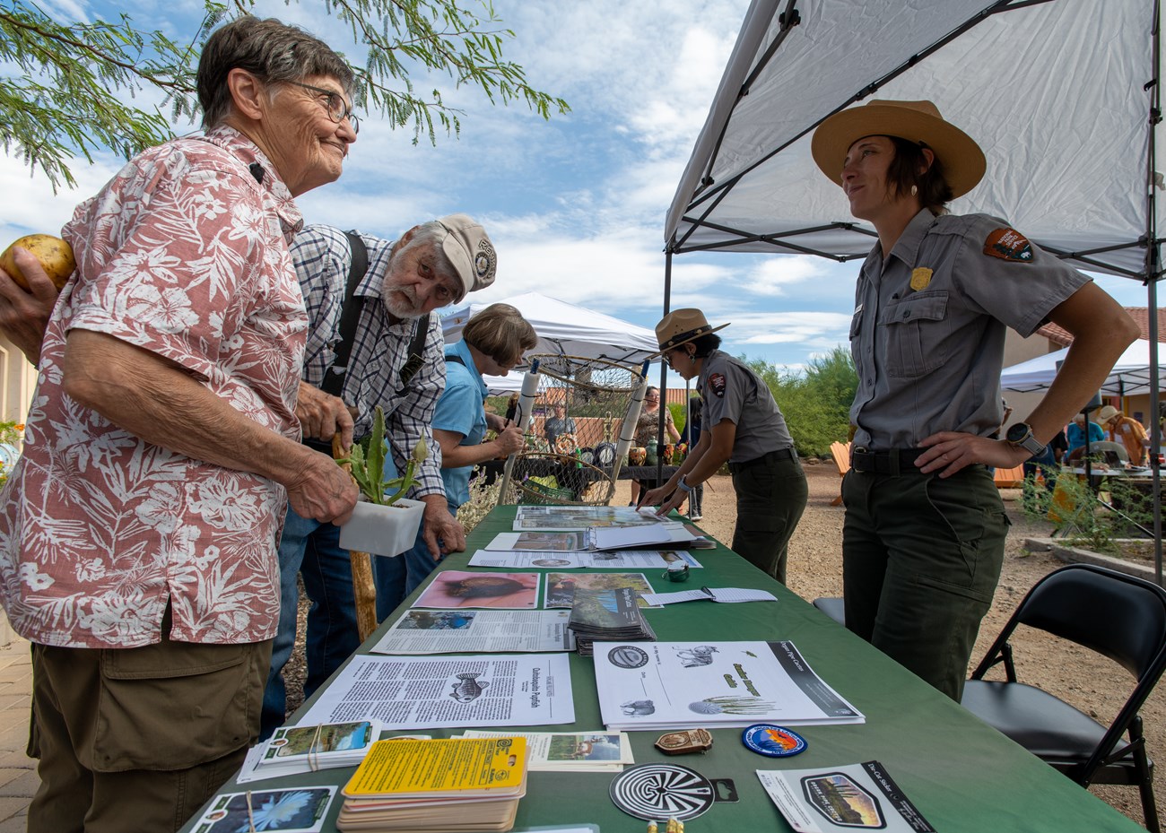 A ranger and a visitor talk across a table outside under a tent.