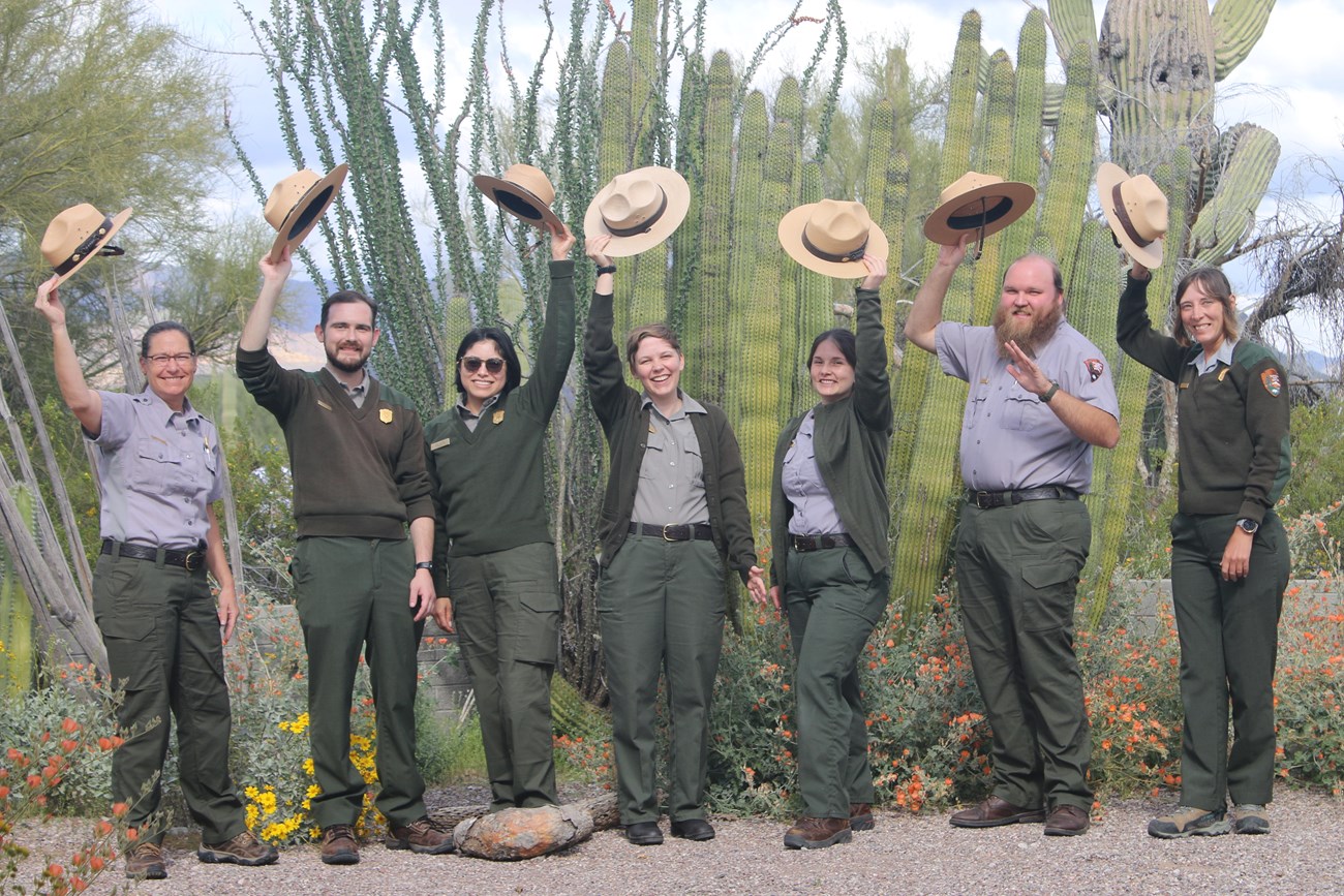 A group of rangers in uniform stand in front of an organ pipe cactus with on hand holding their round flat hats about their heads.