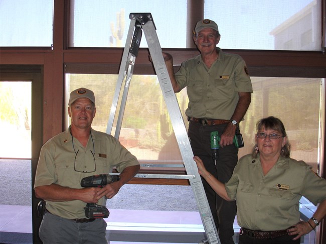 Three volunteers stand together. One on a ladder, two on the ground. One holds a drill.