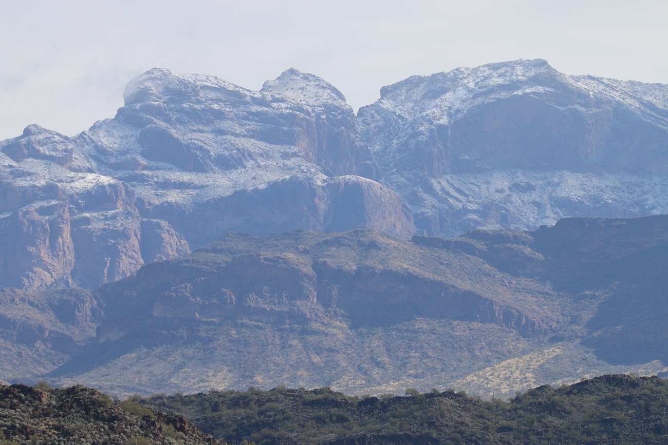 A broad-faced mountain dusted in snow, with a small strip of foreground.