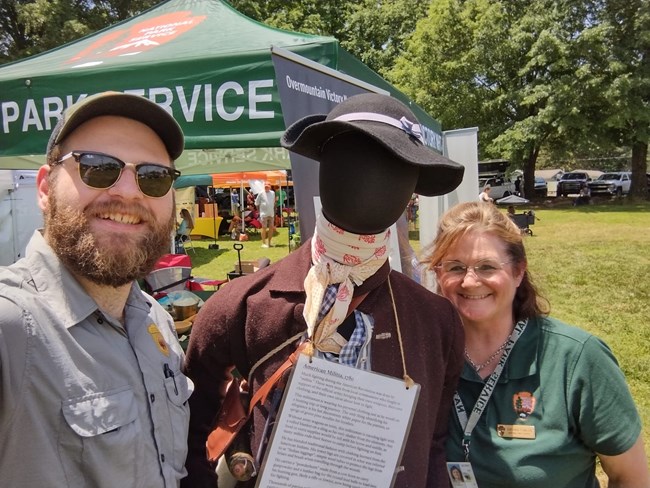 A park ranger and a woman are smiling with a tent and other people in the background.