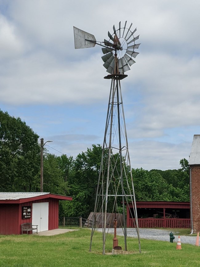 Tall metal windmill that has a "tail fin" behind the blades.  There is a red pump on the ground "inside" the windmill structure.