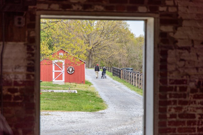 The photograph is taken through a window. Through the window, two visitors can be seen walking along a gravel pathway. One of the visitors is pushing a stroller. To the left of them is a red barn with a brown sign reading "The Story of Grain, Oxon Hill"