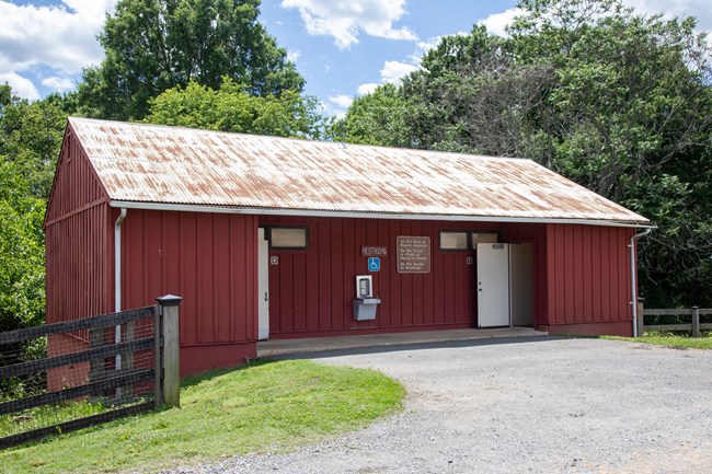 A red rectangular buliding with entrances to men's and women's restrooms and a drinking fountain in the middle next to a wide gravel path.