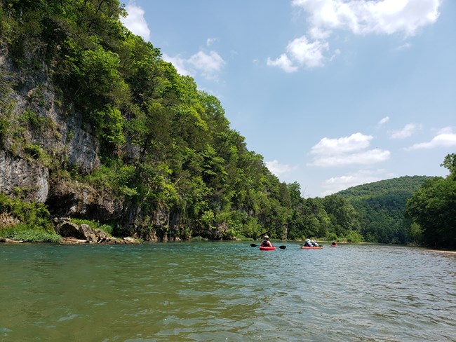 A clear blue river with rock bluffs of dolomite and green trees lining. A group of paddlers in kayaks are headed downstream in front of the camera. A cloudy blue sky is above.