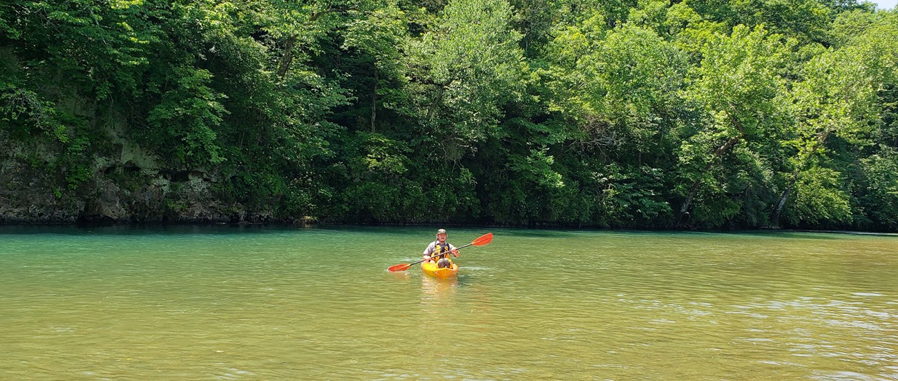A park ranger floats in an orange kayak. The river is calm and blue.