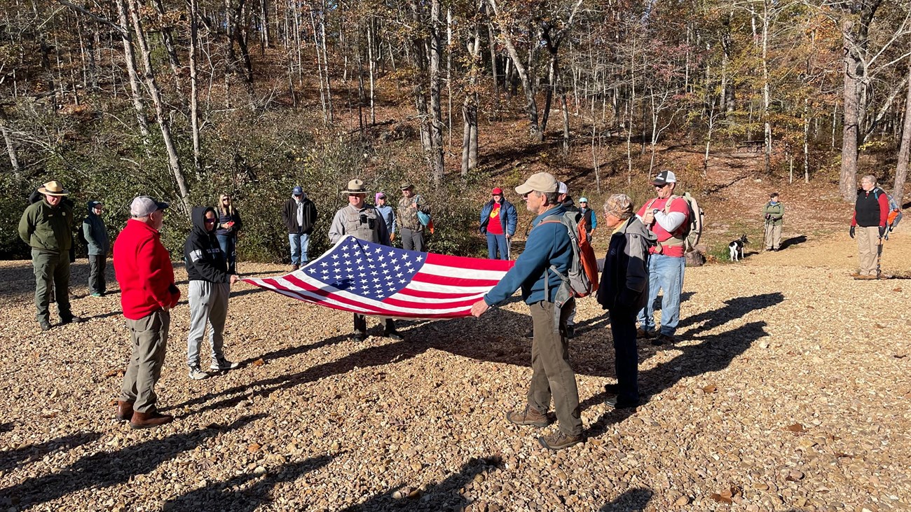 255152350_252938880196559_1314565843731221805_n A group of people gather around a park ranger and veterans holding an American flag on a gravel surface during a ceremony to honor Veterans.