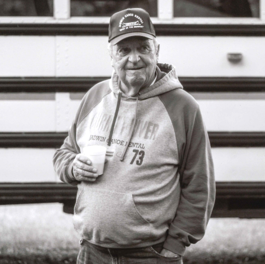 Darrel Blackwell Crop A man stands in front of a school bus with a cup of coffee. His shirt and hat say Jadwin Canoe Rental