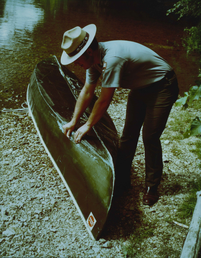 Darrel Blackwell working on a canoe as a Park Ranger From the 70s or 80s: Darrell Blackwell wears a park ranger uniform as he works on a green canoe. He stands on a gravel bar along the edge of the river. The canoe appears to be torn open by a rock, and Darrell inspects the damage.