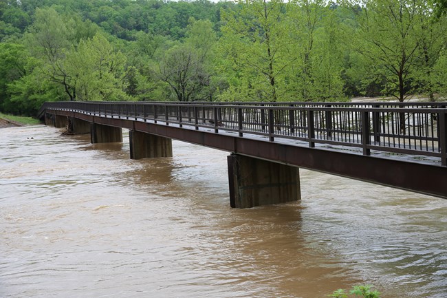A closeup of a brown steel walkway bridge over a small river. The river below is flooding and has brown raging water.