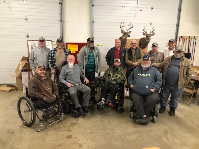 Group of Veterans pose for photo at managed hunt meeting. Some Veterans are standing and some are sitting in mobility devices. Whitetail buck horns mounted on the wall behind them.