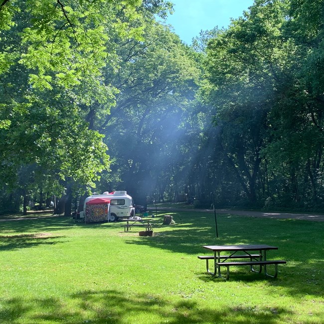 A white camper van sits in a grass field. A smoky haze rises from the campfire.
