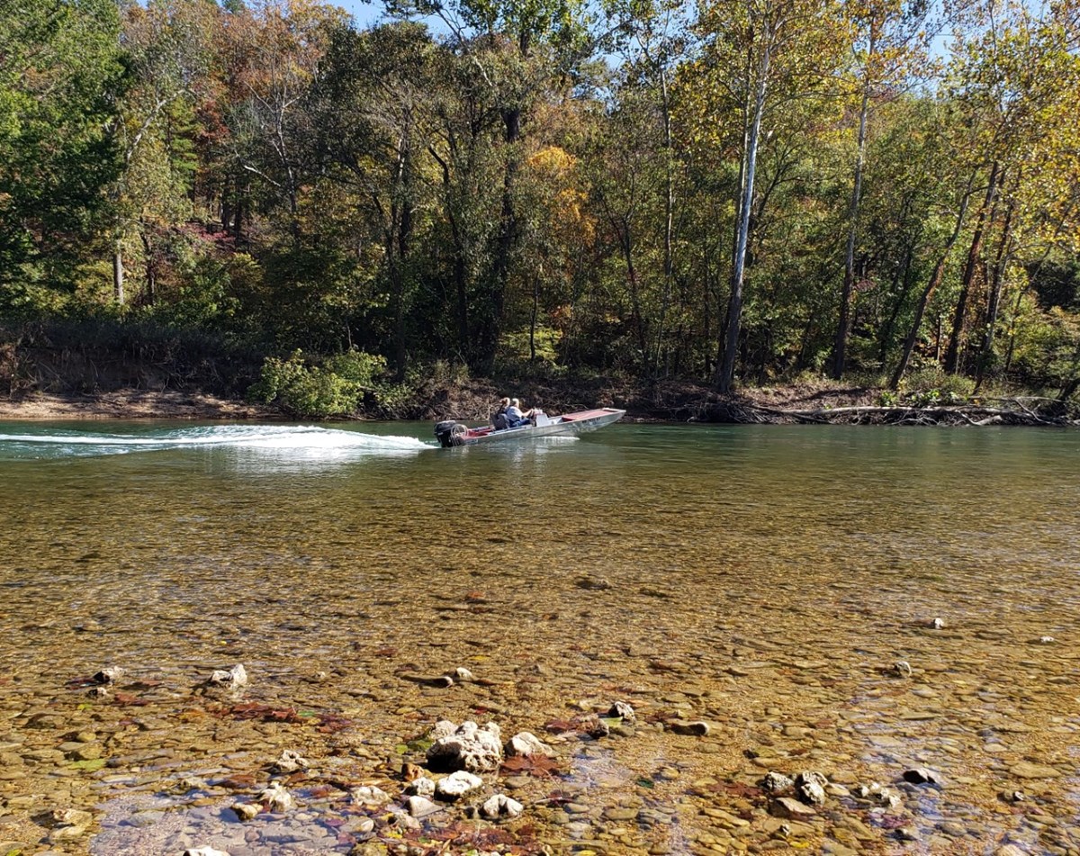 Color photograph looking at a river with gravel bank and forest on the other side. In the water is an aluminum jet boat with two occupants under power headed upstream.