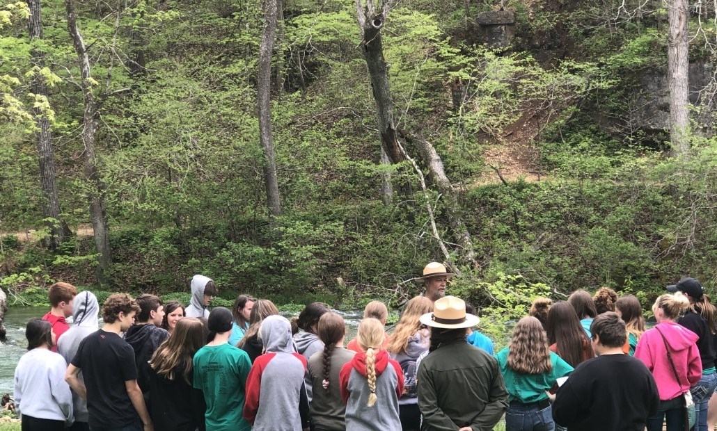 A group of school aged students gather around a park ranger who is presenting next to a running stream.