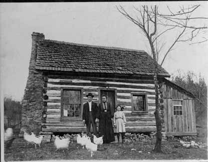 Ozark log house with couple and child in front and chickens in the yard