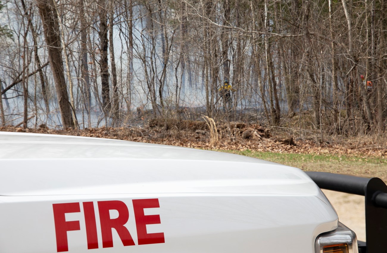 A white fire truck hood up close with words FIRE and a smoky forest fire in the distance looking over the hood.