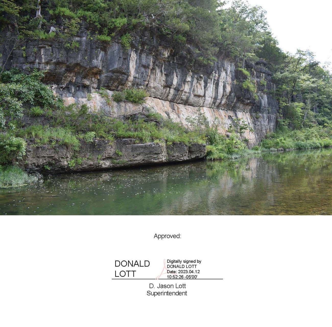 A clear blue stream with a dark rock bluff lining the river. Green vegetation is lining the bluff over the river.