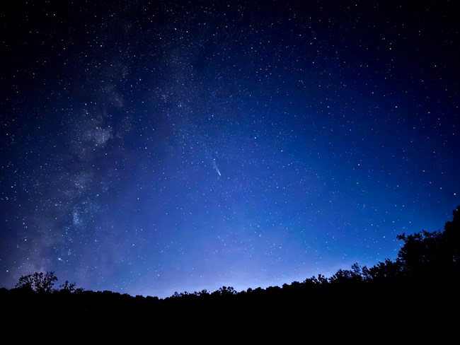 A night sky photo with a comet centered in a field of stars above the horizon. The Milky Way extends upward from the bottom left corner. Lingering skyglow from sunset paints the sky blue-purple.