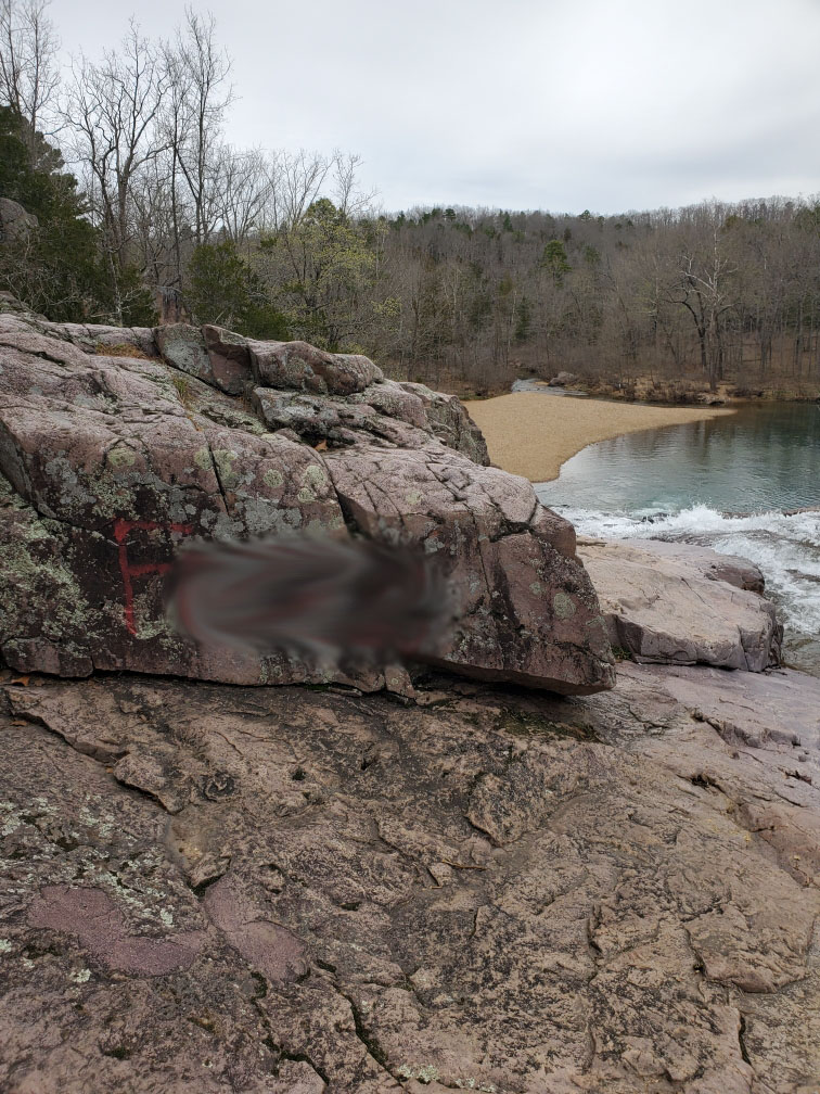 A color photo showing grey rocks along a stream branch with a pool of water below. A red F is painted on the rock face with other letters blurred out.