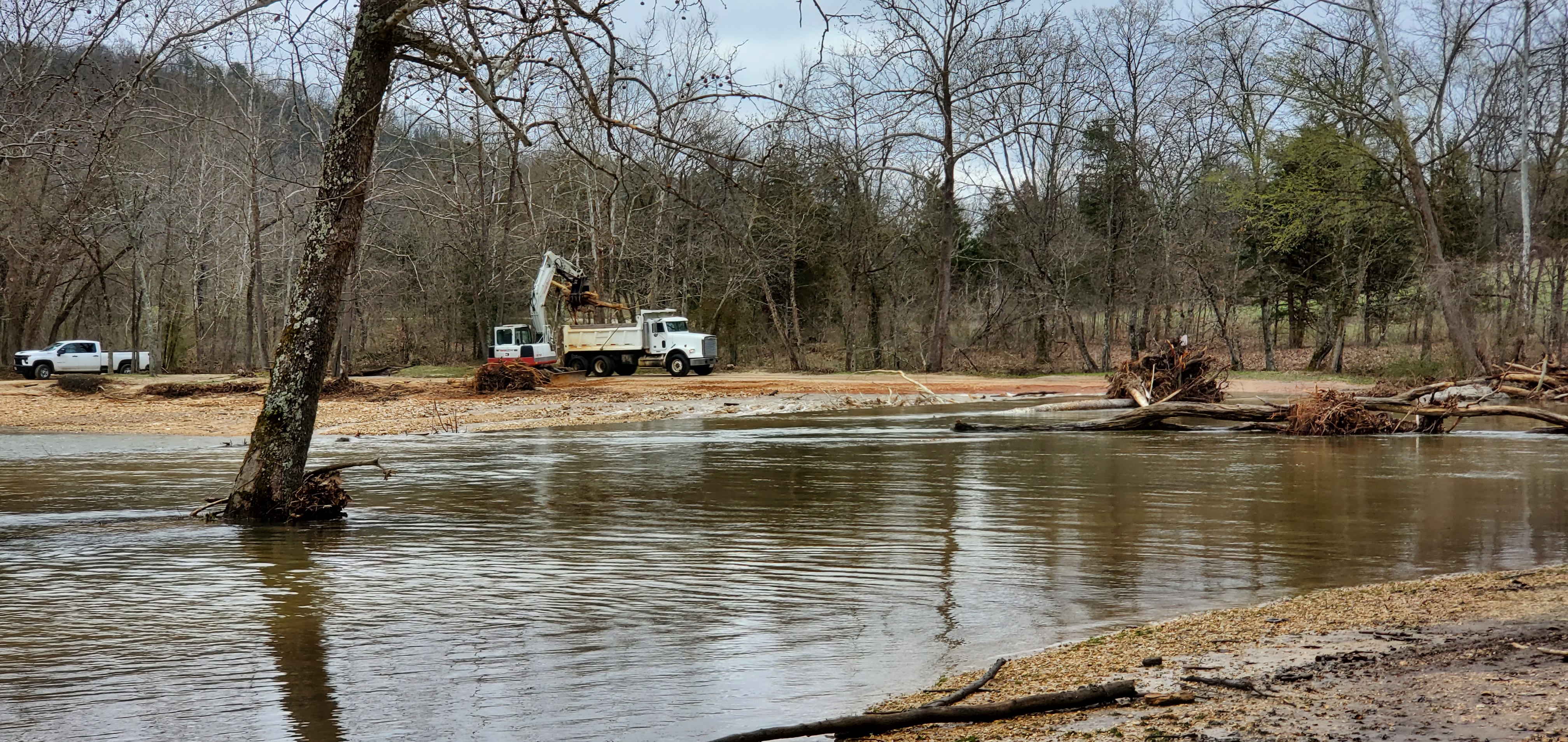 A large dump truck is loaded with fallen trees at a river crossing.