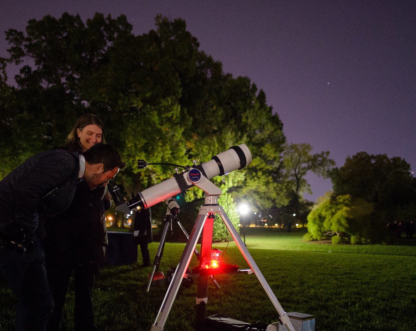 A man and woman look through a white telescope during a night sky event. The area they are in is well manicured park setting with grass and trees. Distant street lights glow in the night sky.