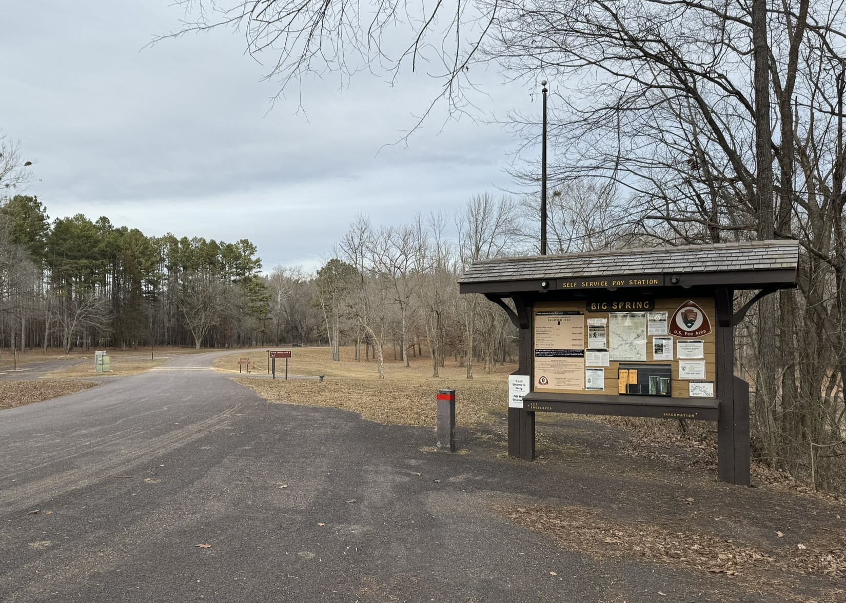 A park bulletin board covered with literature, sign says self service pay station, Big Spring. Asphault campground and other signs can be seen in distance. Pine trees are lining the roadway.