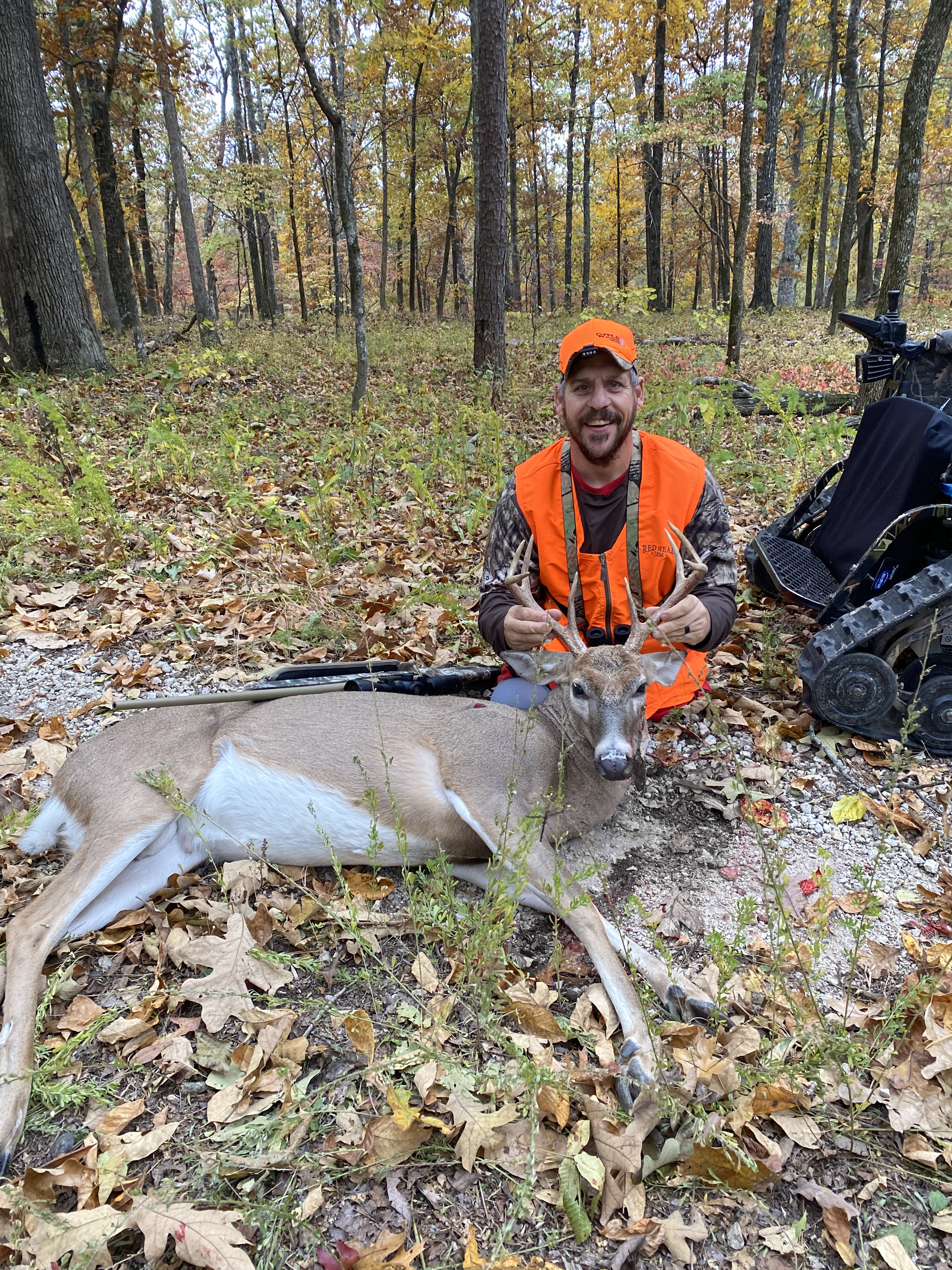 Another successful hunt! A man in a hunter orange outfit smiles at the camera. A freshly-hunted deer lies in front of him.