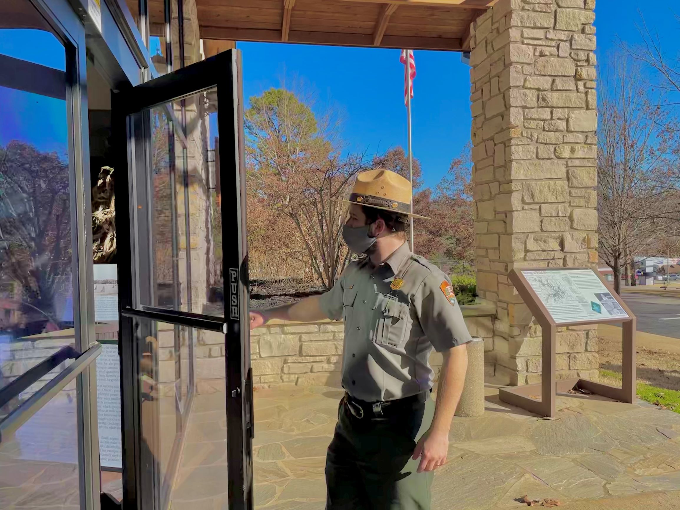 A male park ranger in uniform and flat hat wearing a mask entering into a building through the glass door. A wayside is in distance next to a light brown stone pillar in the entranceway.