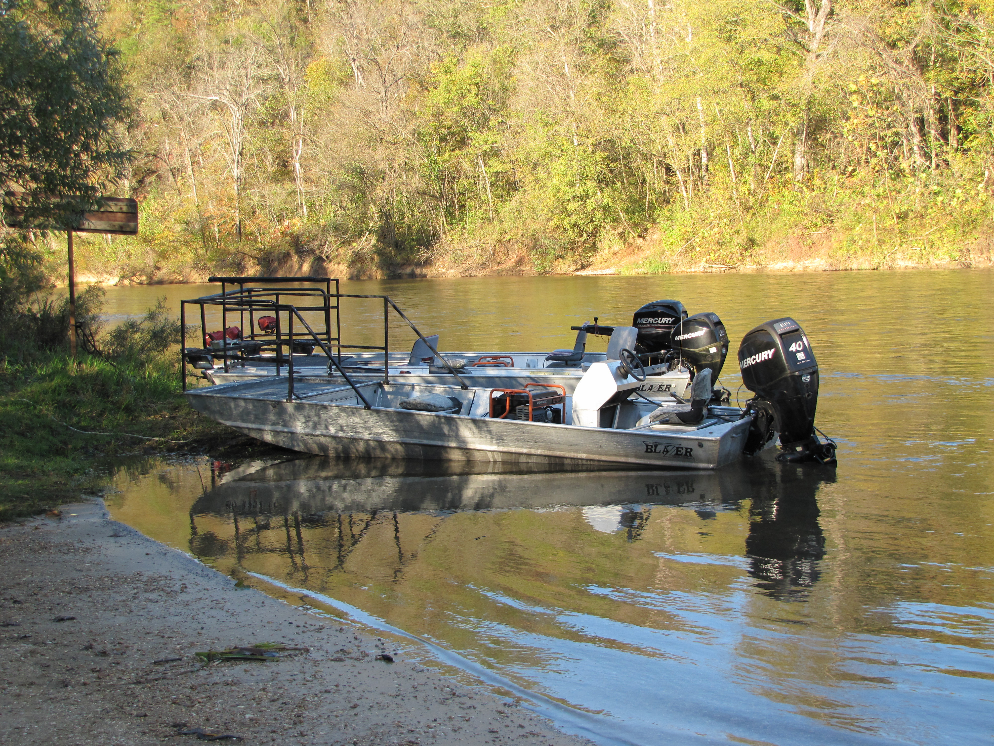 3 aluminum flat bottom jet boats are on the edge of the river during the fall. a generator is in the boat with gigging rails on the front.