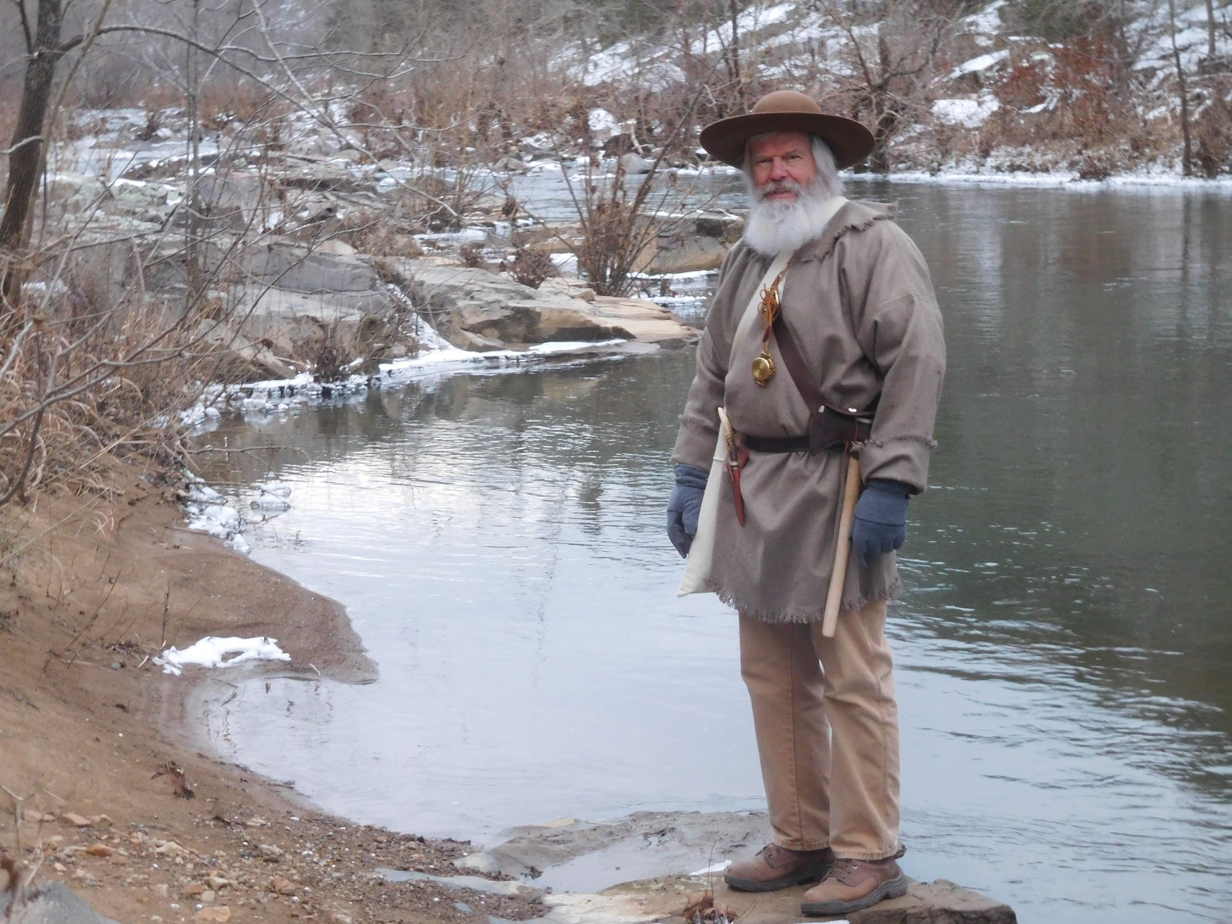 man dressed in 1700s trapping clothing with hat and white beard standing by a snow covered river