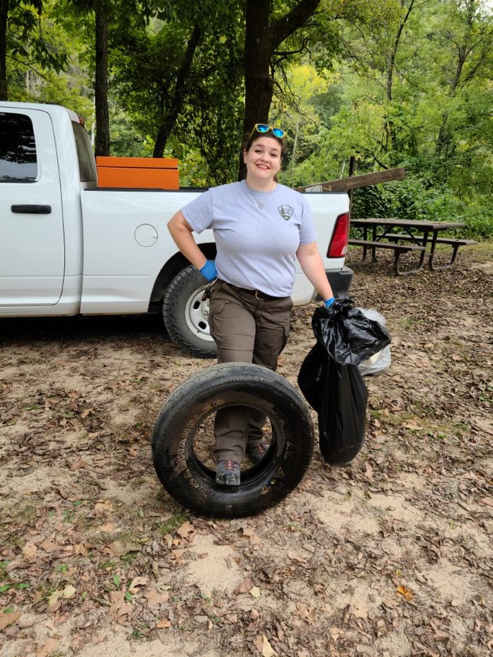 Pulltite cleanup A park service employee in grey NPS t-shirt and green pants holding a trash bag and resting foot against a car tire during a cleanup.