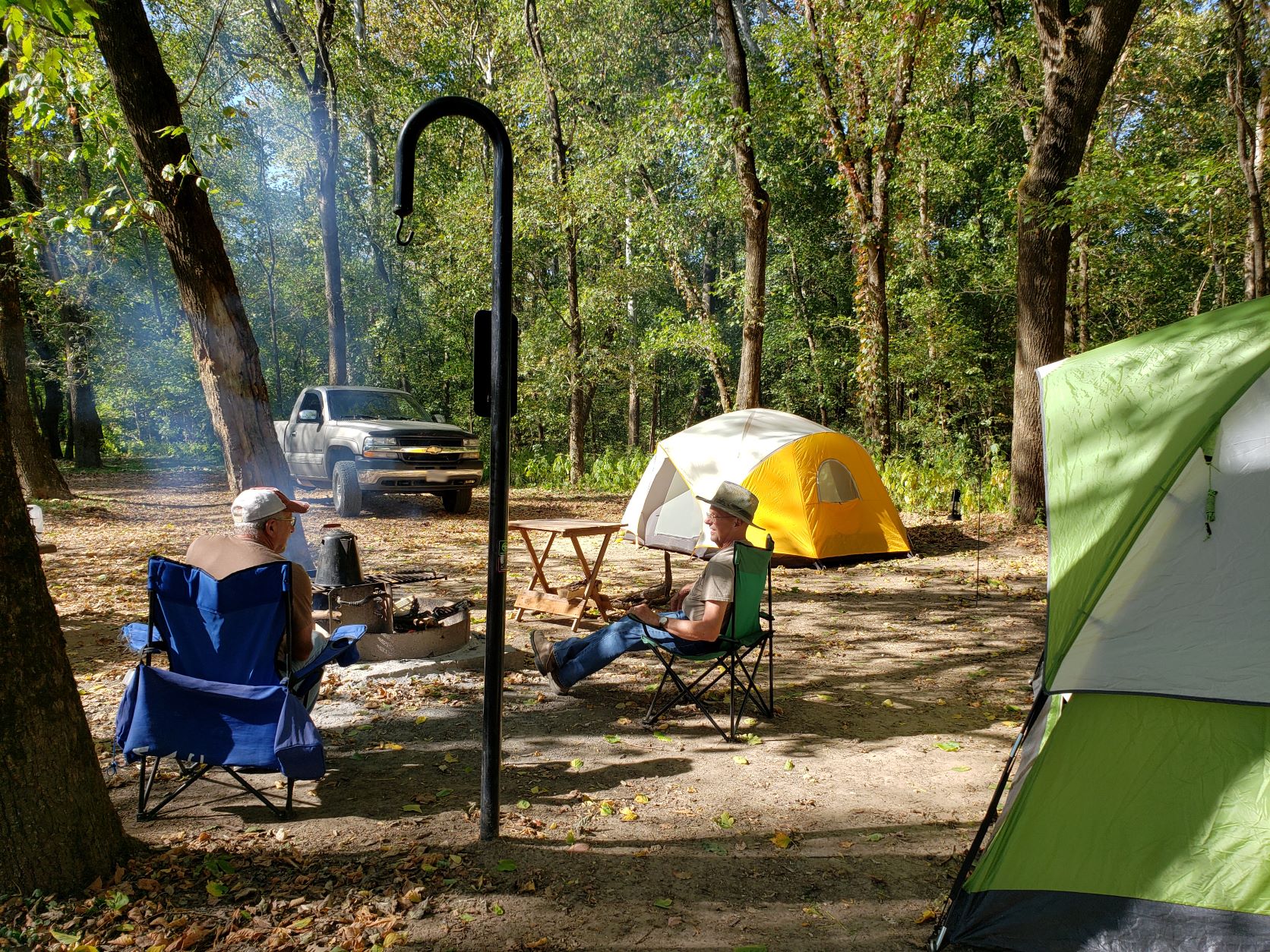 two gentlemen sit around a campfire during the day, tents and a truck are visible