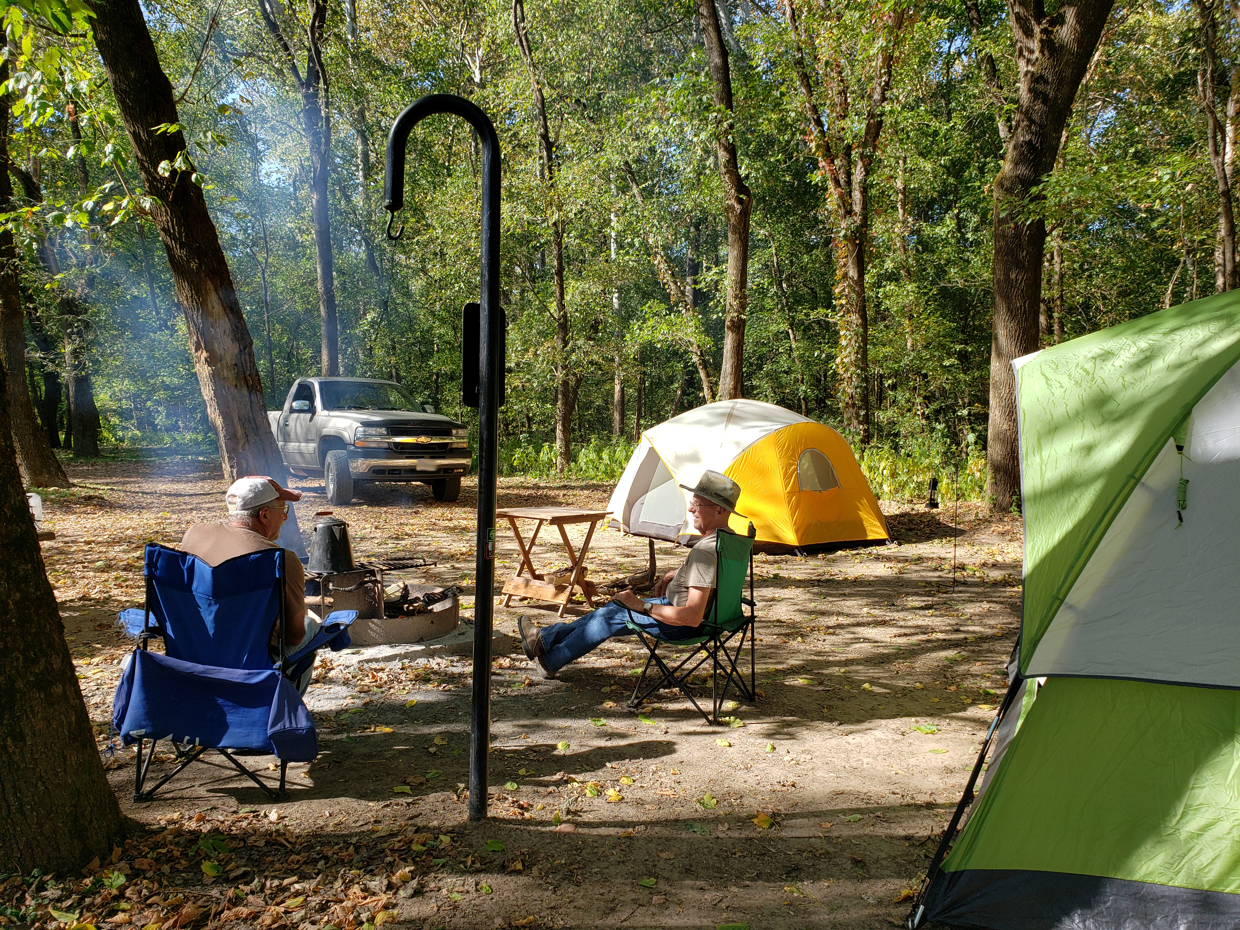 A campsite with tents and people in lawn chairs around a fire ring and truck in distance.