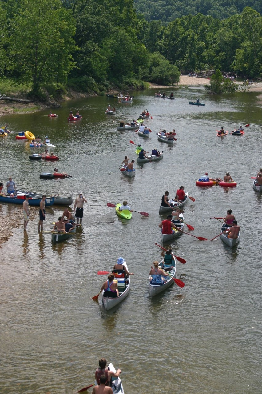 canoes and kayaks going down a river with green trees lining the bank