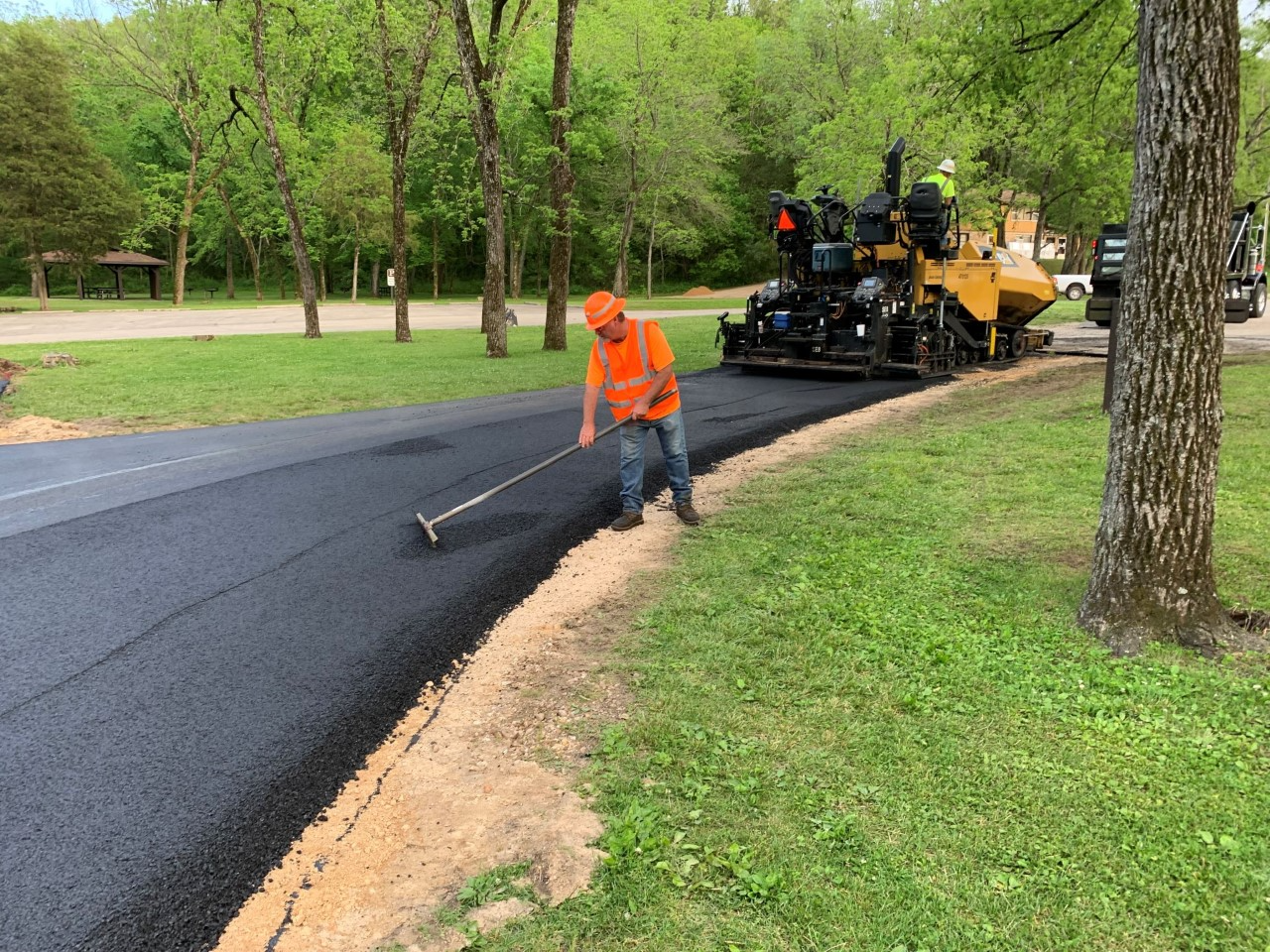 A road worker in bright orange safety vest is working asphalt with a rake on a roadway in a campground with a paver in the background