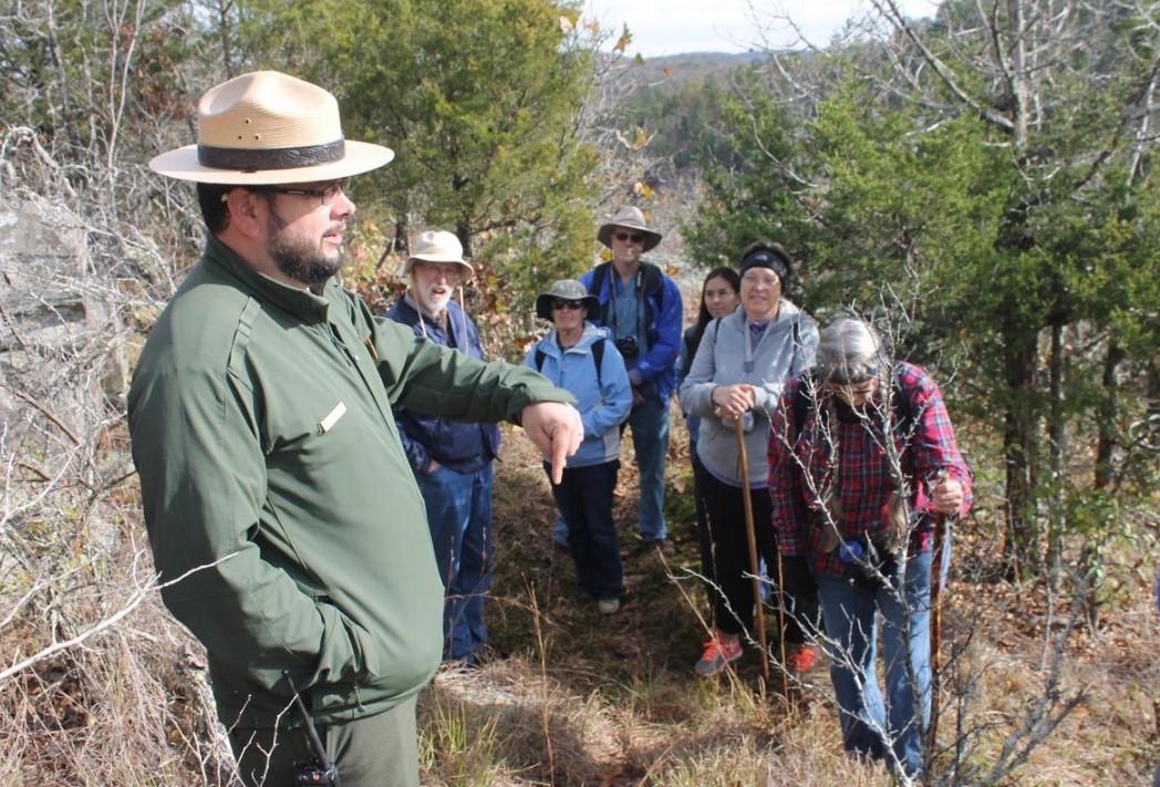 A park ranger in uniform talking to a group of hikers with foliage in the background