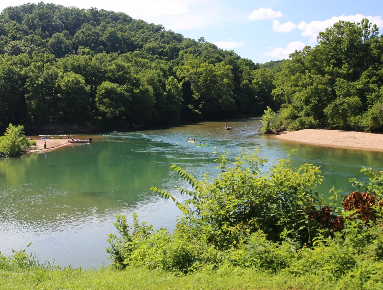 A river scene with rolling hills and gravel bars. kayaks and john boats are in the river.