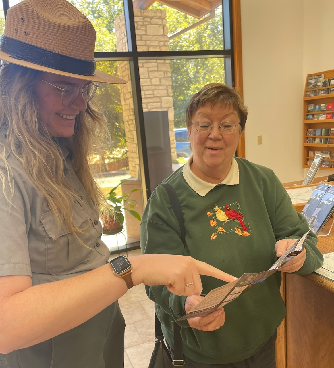 downloaddchgfdhgfdhgf A close up of a park ranger helping a visitor in the lobby of a visitor center. The two visitors both have a smile on their face. The park ranger is on the left and the woman visitor on the right. Clear large windows are seen behind them.