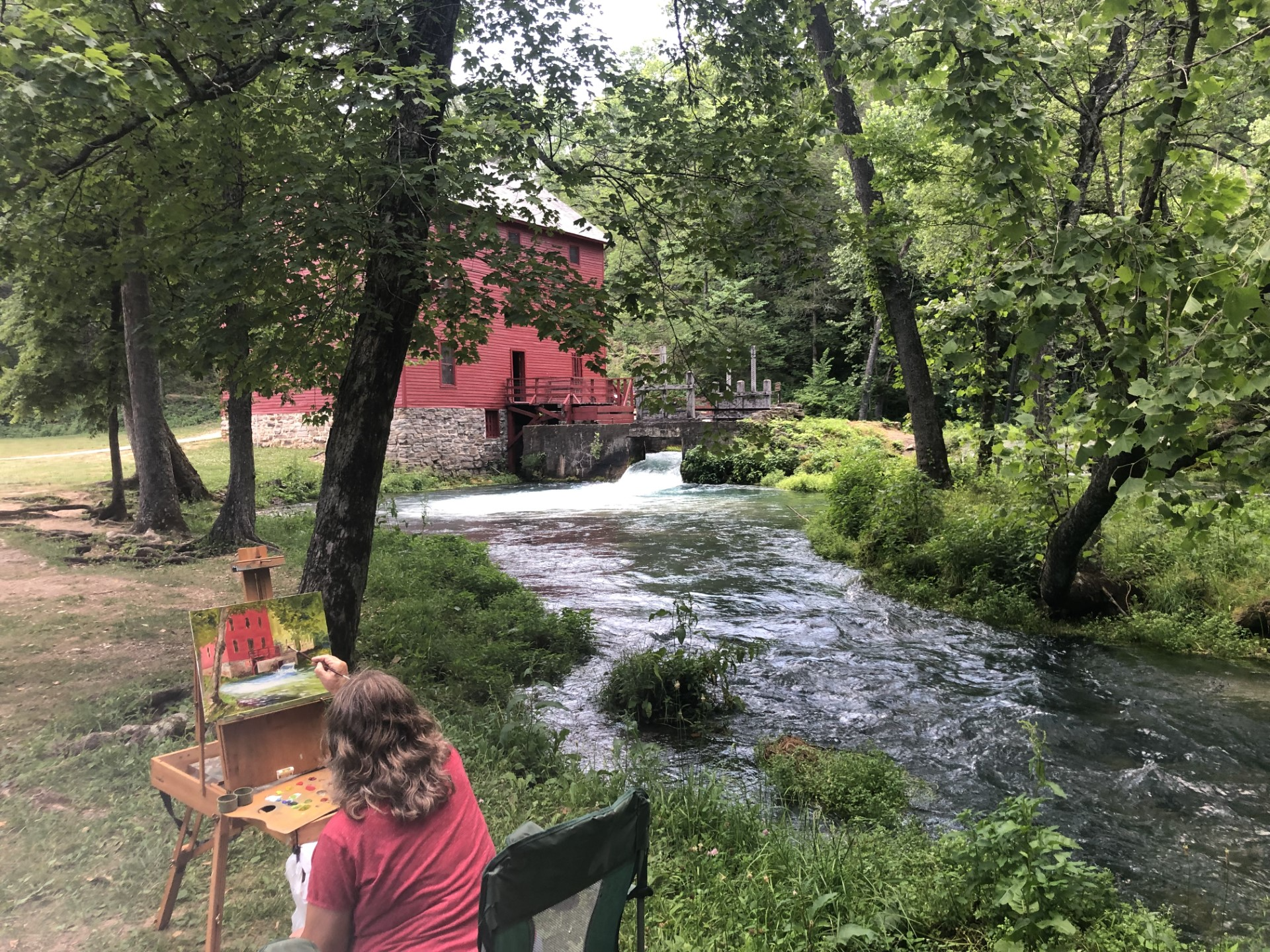 download//// A woman paints alley mill on a canvas while in front of the red alley mill and blue spring.