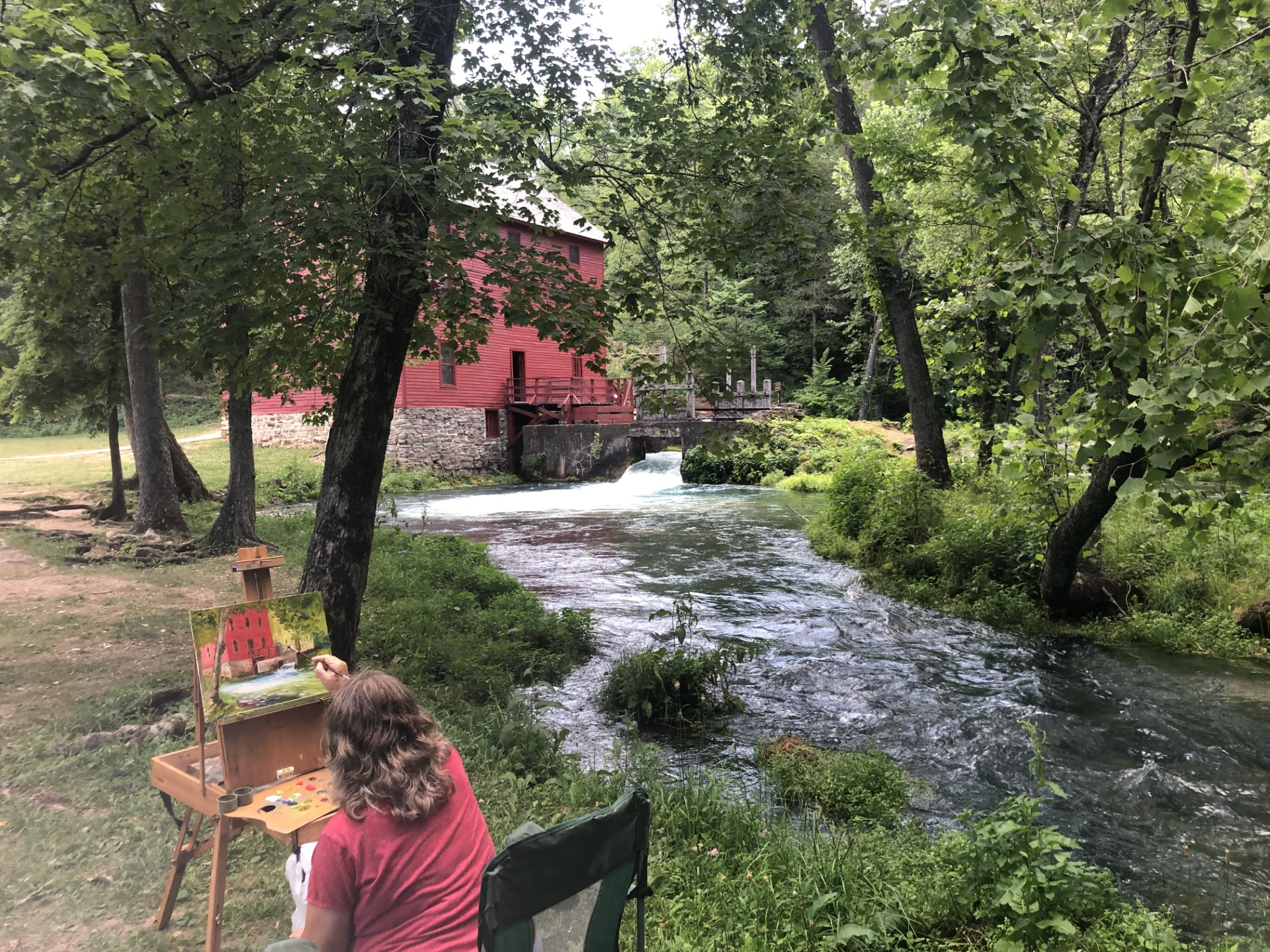 A woman paints alley mill on a canvas while in front of the red alley mill and blue spring.