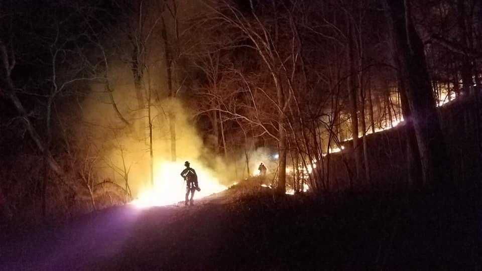 a wildfire burns on a hill at night with firefigher in foreground