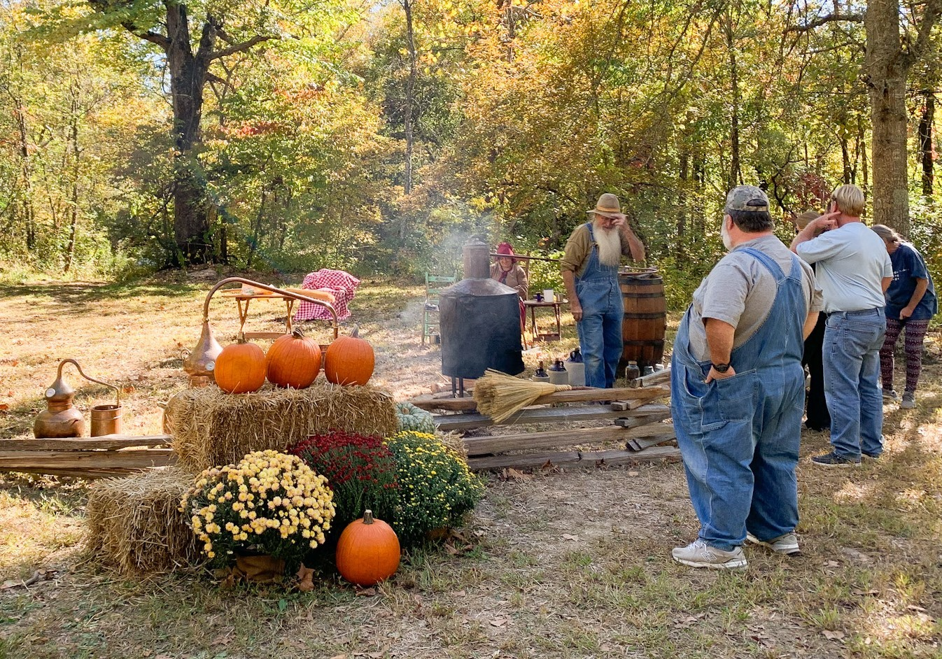 downloahhd A fall scene with changing leaves. A man and woman dressed in costume of Ozark Pioneers are demonstrating moonshine and broom making. Pumpkins and mums are in front of a split rail fence.