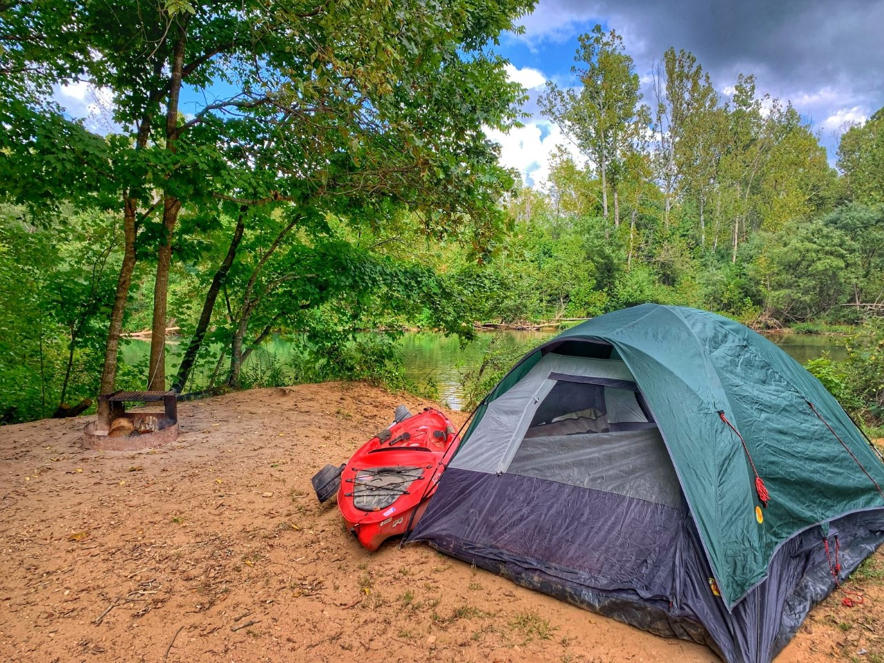 fsdfasfed A campsite by river with green tent, red canoe by tent, fire ring and river in the background. The camp is empty.