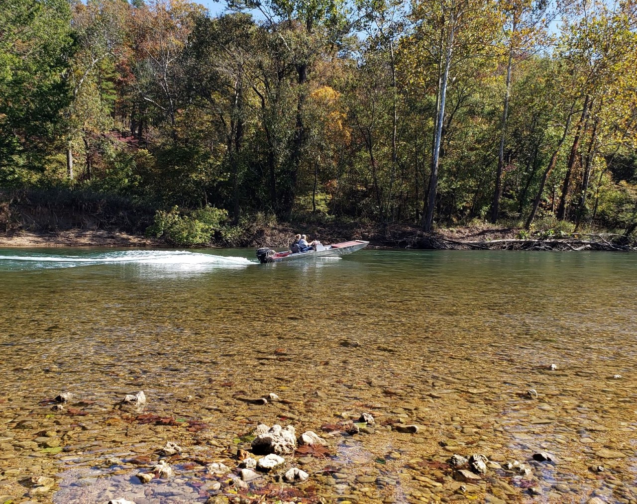A flat bottom jet boat under power runs up a small clear stream with two occupants.