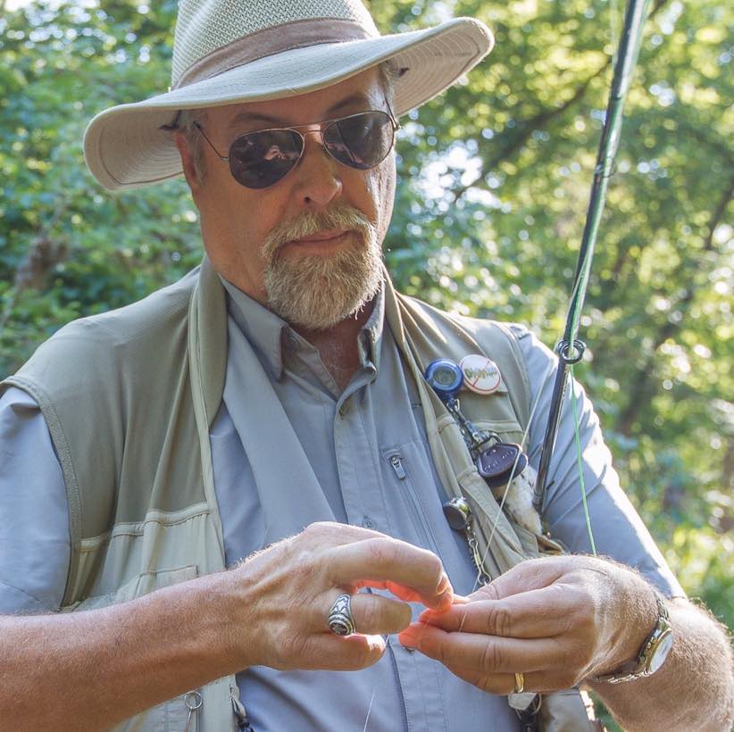 A fisherman with hat and fishing vest holds a fly rod and is looking down at his lure