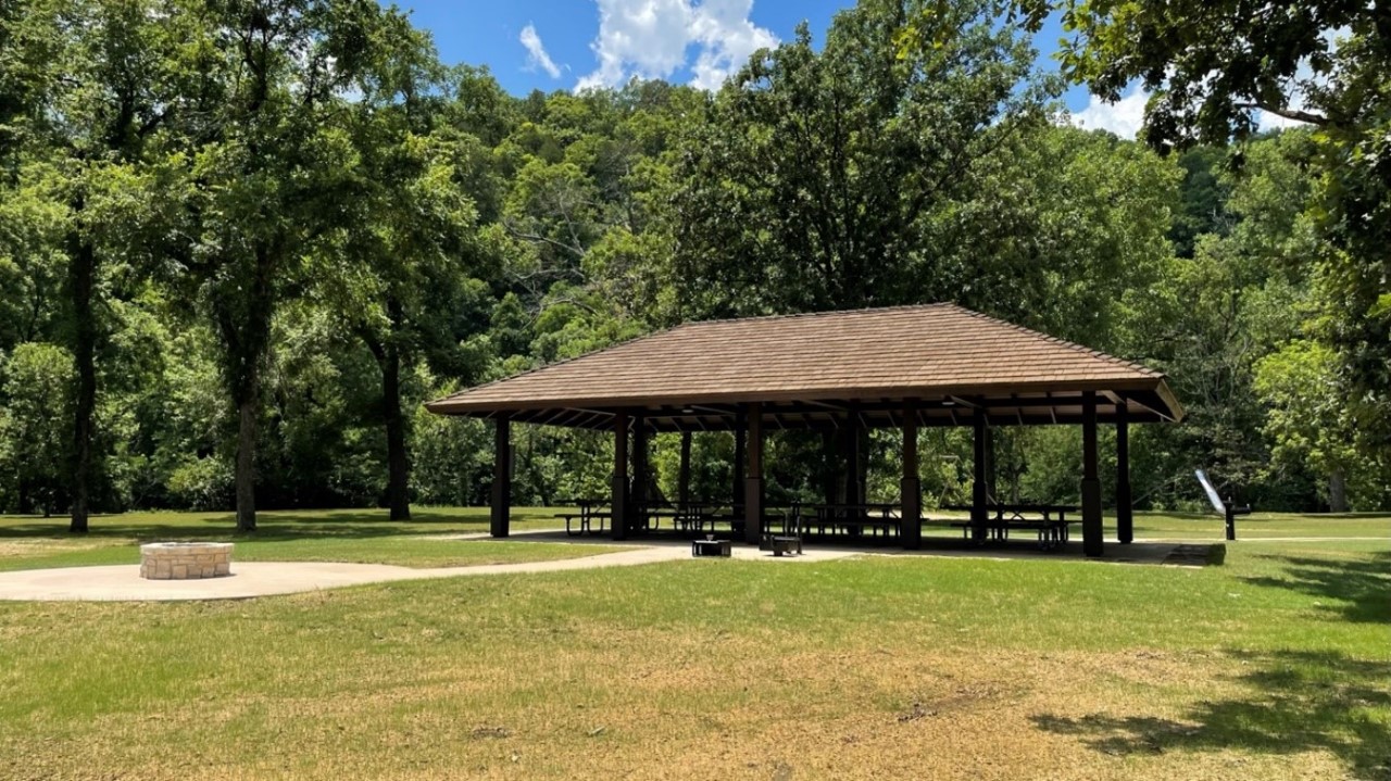 thumbnail_Big Spring pavilion A brown park pavilion in a grassy area with a green forest behind it and a clear blue sky. The pavilion is empty of people but picnic tables are seen inside it.