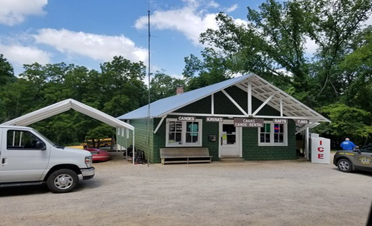 thumbnail_Carrs Store (1) A green building with aluminum roof and white trim. Text on building says Carrs Canoe Rental. Four other wooden signs on building say Canoes, Kayaks, Rafts, Tubes. Two vehicles are in the gravel parking lot.