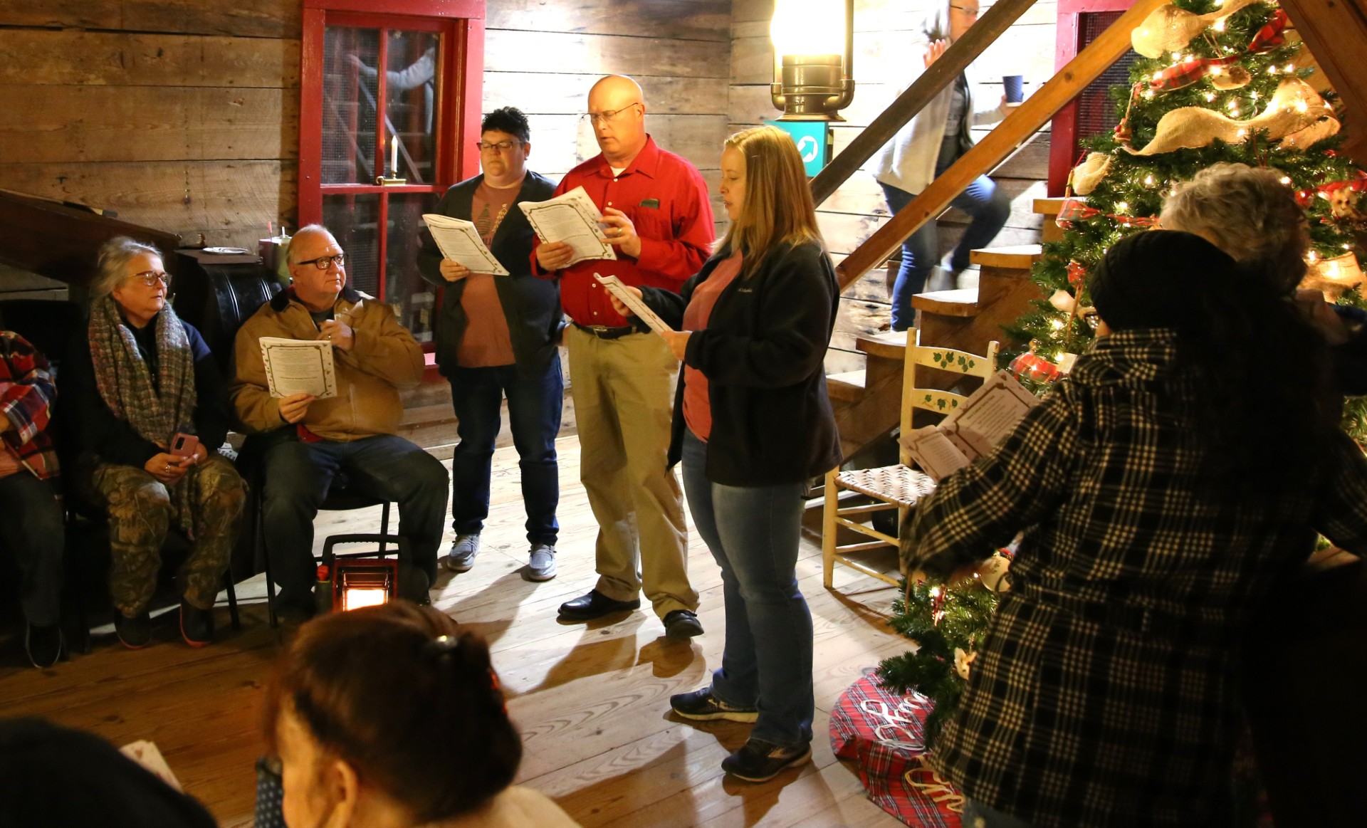 A group of carolers gathered in an old mill singing by candlelight. A Christmas tree is in the corner near the singers.