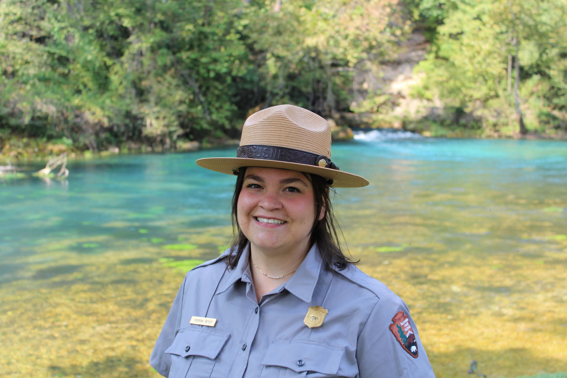 thumbnail_IMG_5595edit A female park ranger in flat hat and national park service uniform. A clear blue spring is in the background.