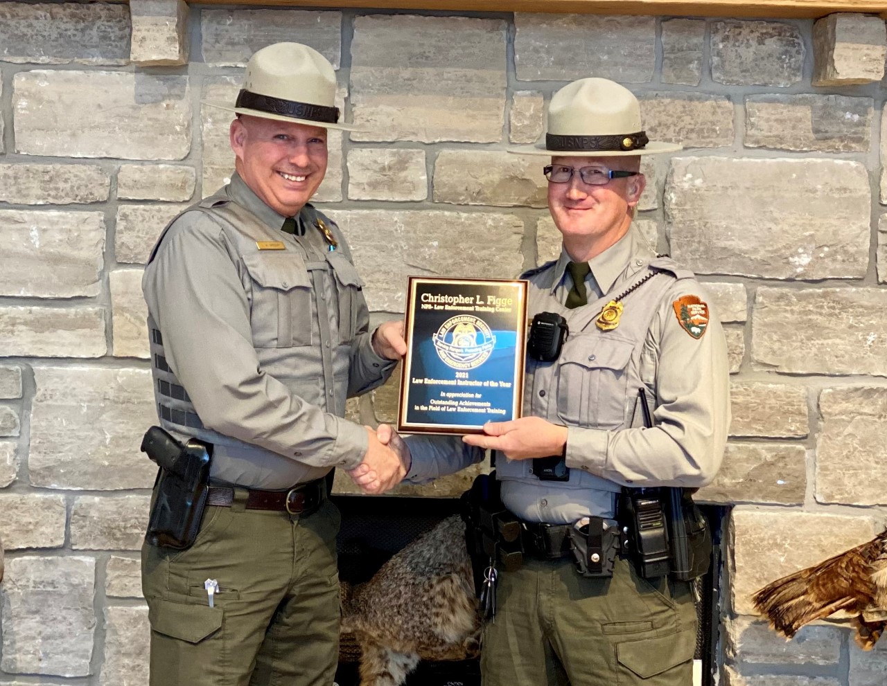 Two National Park Service law enforcement rangers in flat hats and gear standing in a lobby holding an award plaque between them.